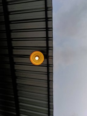 A light bulb of a lamp light hanging under a galvanized steel roof. Also the background is a cloudy sky clipart