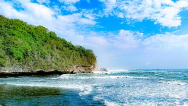 Arka planda büyük bir kaya oluşumu olan kayalık bir kıyı şeridi. Su sakin ve gökyüzü mavi.
