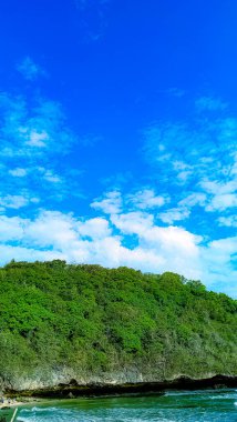 Arka planda büyük bir kaya oluşumu olan kayalık bir kıyı şeridi. Su sakin ve gökyüzü mavi.