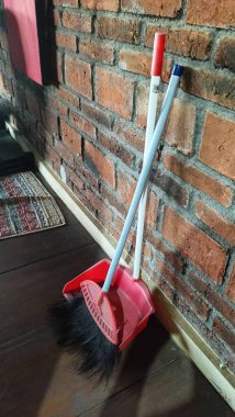 Kırmızı bir faraş ve süpürge kırsal bir tuğla duvara yaslanmış, rahat ve pratik bir temizlik düzeneği yaratıyor. Oda servisi ve temizlik için mükemmel..