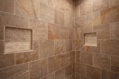 A modern shower with stone tile walls featuring built-in shelves. Surfaces showcase a natural stone look with varied earthy tones and textures. clipart