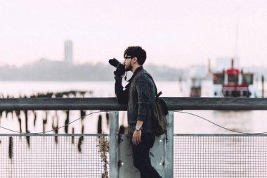 Sırt çantalı bir fotoğrafçı şehir manzarasını yakalamak için kamera kullanır. Nehir kenarındaki bir rıhtımda durur ve doğa ile şehir yaşamının karışımını yansıtır..