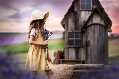 Fotoğrafta açık yaz elbisesi ve çiçekli hasır şapka giymiş bir kız görülüyor. Elinde bir demet lavanta tutuyor ve arka planda eski ahşap bir eve bakıyor.