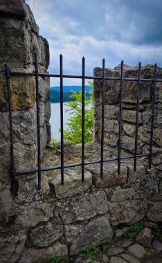 The photo was taken through an ancient stone window reinforced with iron bars. Views from the window open to the lake and forested mountains under a cloudy sky. clipart