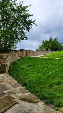 Fotoğraf yeşil otlar ve ağaçlarla çevrili bir tepeye uzanan dolambaçlı bir taş patikayı gösteriyor. Bulutlu gökyüzü sakin bir atmosfer yaratır