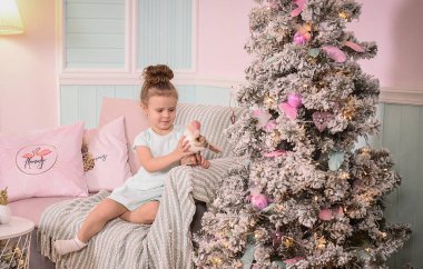 Fotoğrafta güzel bir saç stili ve açık renk elbisesi olan bir kız var. Süslü bir Noel ağacı, ışıklar ve Noel atmosferinin yanında..