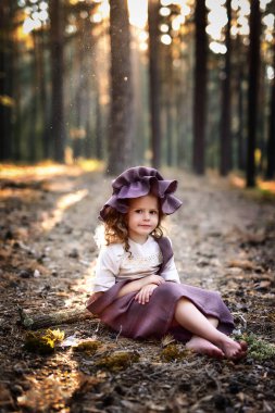 Bu, yüzünde hafif bir gülümsemeyle ormanda yerde oturan bir kızın görüntüsü. Şirin bir elbise ve şapka giyiyor.