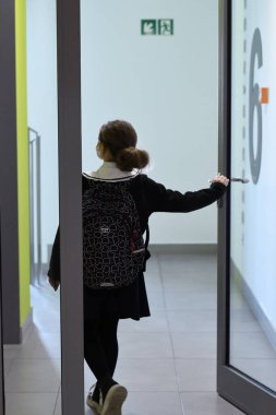 Okul üniformalı bir kız camdan kapıdan girişe doğru geliyor. Üzerinde desenli siyah bir sırt çantası yama yakalı beyaz bir bluz ve siyah tayt var..