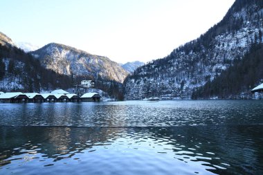 winter landscape with a lake and mountainous shores covered with snow. Wooden buildings are visible in the foreground clipart