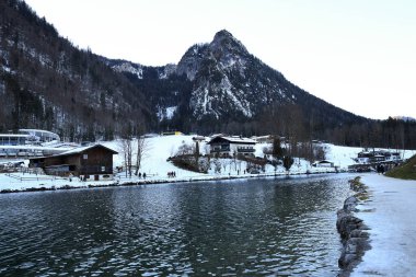 winter landscape with a lake and mountainous shores covered with snow. Wooden buildings are visible in the foreground clipart