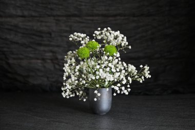 Siyah desenli arka planda yer alan küçük gri bir vazoda çingene çiçekleri ve yeşil kasımpatı kompozisyonu