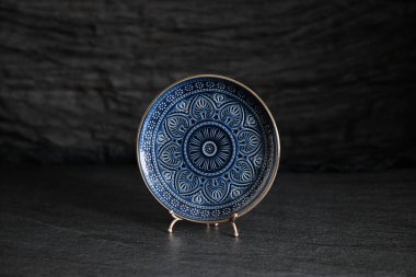 Ortasında bir süs bulunan mavi bir plaka tasvir eder, koyu dokulu bir arka plana karşı bir stantta yer alır.