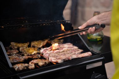 Sarı tişörtlü birinin arka bahçede ızgaradan yükselen dumanla açık bir barbeküde elinde maşayla et kızarttığını yakın.