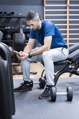 Odaklanmış genç bir adam spor salonundaki bir bankta dinleniyor, antrenman sırasında telefonunu kaydırıyor.
