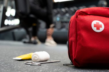 Tıbbi malzemelerle dolu kırmızı bir ilk yardım çantası spor salonunun zemininde otururken arka plandaki kişi yaralı bacağını tutuyor.