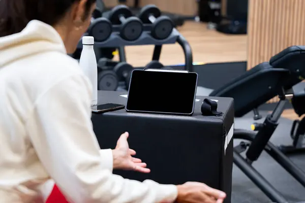 Spor kıyafetleri içinde bir kadın spor salonunda oturup, bir tablet ekranı izliyor ve çevrimiçi fitness antrenmanına hazırlanıyor.
