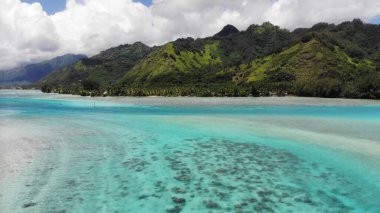 Mercan resifleri ve bulutlu tropikal gökyüzünün altındaki yemyeşil dağlarla turkuaz bir lagünün çarpıcı manzarası.