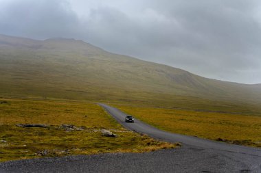 İzlanda 'da sisli kayalar ve dramatik İskandinav arazisiyle çevrili sessiz bir dağ yolundan geçen siyah bir araç görülüyor.