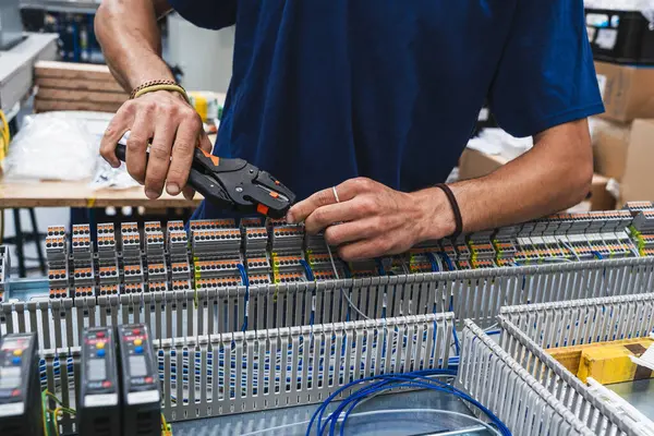 Endüstriyel elektrik kontrol paneli üzerinde çalışan teknisyenlerin yakın çekimi. Görüntü, mühendislik, elektrik, bakım ve otomasyon gibi kavramları temsil eden kablolar ve elektronik bileşenlerle yapılan elle yapılan hassas çalışmaları gösteriyor..