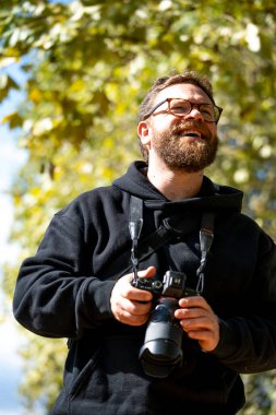 A bearded man wearing glasses and a black hoodie is reviewing his camera screen indoors surrounded by soft natural light creating an intimate and focused atmosphere with a clear emphasis on concentration and creativity