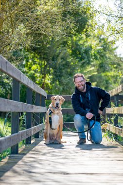 A bearded man walking with his dog on a peaceful forest trail wearing casual clothes and camera gear the path is surrounded by green trees and bathed in soft sunlight on a quiet nature walk