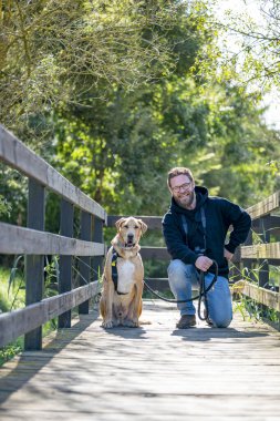 A bearded man walking with his dog on a peaceful forest trail wearing casual clothes and camera gear the path is surrounded by green trees and bathed in soft sunlight on a quiet nature walk