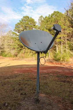 A weathered satellite dish on a metal pole, surrounded by a grassy yard and red soil, with a backdrop of trees under a partly cloudy sky. clipart