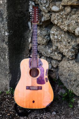 Eski akustik gitar, kırsal bir ortamda, güpegündüz engebeli bir taş duvara karşı duruyor.