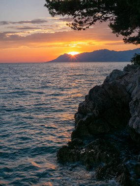 Altın güneş ışığı uzak dağların ardında batarken sudan yansıyarak kayalık kıyı boyunca sakin bir atmosfer yaratır. Gökyüzü alacakaranlıkta sıcak renklerle boyanır..