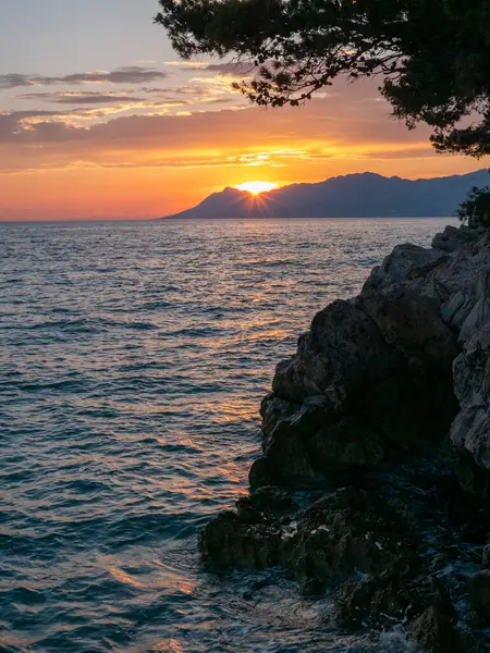 Altın güneş ışığı uzak dağların ardında batarken sudan yansıyarak kayalık kıyı boyunca sakin bir atmosfer yaratır. Gökyüzü alacakaranlıkta sıcak renklerle boyanır..
