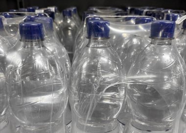 Temiz plastik su şişeleri ambalajda düzgünce düzenlenir, mavi kapaklarının altını çizer. Dekor bir market koridoru, satılık ürünleri sergiliyor..