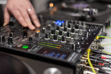 Bir el DJ kurulumunun karıştırma konsolunu çeşitli düğmeler ve kaydırma çubukları ile ayarlar. Müzik havayı doldururken mekan heyecanla uğulduyor, heyecan verici bir hava yaratıyor..