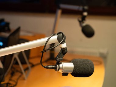 İyi donanımlı bir yayın stüdyosunda tezgahta bir mikrofonun yakın çekimi vardır. Kayıt oturumu hazırlıkları devam ederken, yumuşak ışıklandırma odaklı atmosferi güçlendiriyor.