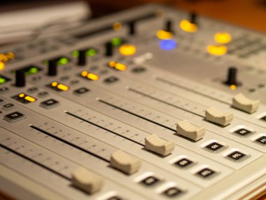 Bir müzik stüdyosunda çeşitli sürgülü ve aydınlatılmış düğmelerin yer aldığı bir karıştırma konsolu bulunmaktadır. Cihazlar, odaklanmış aktivite ve canlı ışıklarla aktif bir kayıt oturumu olduğunu gösteriyor..