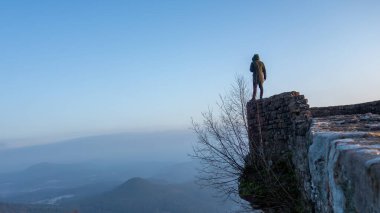 Yeşil ceketli yalnız bir figür yıpranmış bir taş duvarın kenarından uzak dağ manzaralarına bakıyor. Sakin sabah atmosferi yumuşak mavi bir gökyüzü tarafından güçlendirilir..