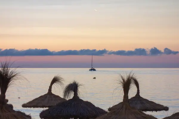 Bir yelkenli gün batımında sakin sularda zarifçe hareket eder ve ön planda plaj şemsiyeleri görünür hale gelir. Gökyüzü yumuşak pastel renklerle boyanmıştır..