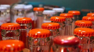 Canlı turuncu başlıklı cam şişe koleksiyonu bir pazar masasında düzenlenir. Bu canlı ekran güneşli bir açık hava ortamında, serinletici seçenekler sunar.