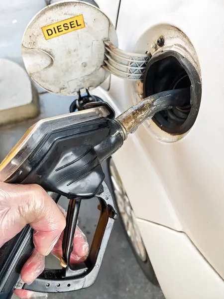 Güneş ışığı altında benzin istasyonunda beyaz bir araca mazot doldururken bir el hortumu tutuyor. Yakıt kapağı açık ve çevredeki alan hareketli görünüyor..