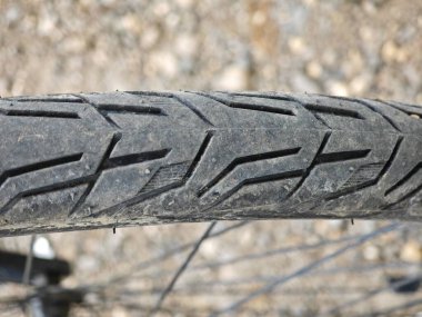 Bu yakın plan, çakıllı bir yolda dinlenen bisiklet lastiğinin lastik izlerini yakalar, desenli yüzeyi ve aşınmış yivleri gösterir. Açık mavi gökyüzü parlak güneş ışığını yansıtır.