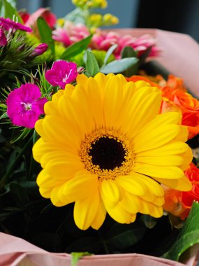 Canlı sarı bir gerbera papatyası, doğanın güzelliğini ve çeşitliliğini gösteren, çeşitli renklerdeki çiçeklerle dolu, yemyeşil bir bukette dikkatleri çeker..