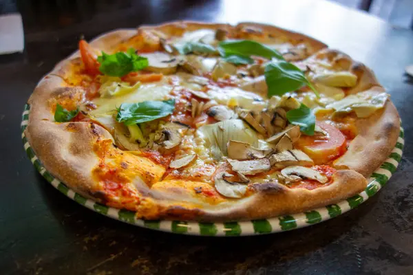 Yeni pişmiş bir vejetaryen pizzada mantar, domates, soğan ve şifalı otlar dahil olmak üzere çeşitli malzemeler yer alır. Tabak, rahat bir restoranda çizgili yeşil ve beyaz bir tabakta servis edilir..