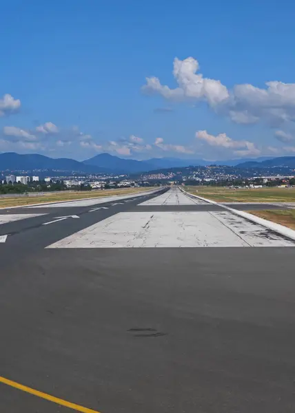 Uzun bir havaalanı pisti parlak mavi bir gökyüzünün altında ufka doğru gidiyor. Çevredeki binalar manzarayı işaretlerken, arka planda dağlar görülebilir. Atmosfer sakin ve davetkar..