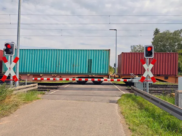 Kırmızı ışıklar ve indirilmiş bariyerler trafik tıkanırken bir yük treninin kırsal bir geçitten geçtiğini gösteriyor. Açık gökyüzü öğleden sonraki ayarı güçlendirir.