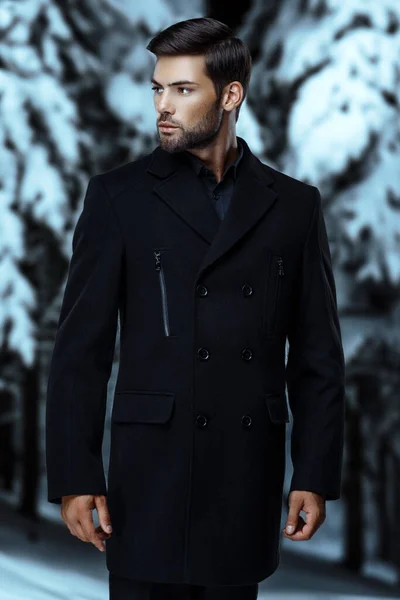 handsome sexy man with a fashionable haircut in a winter black coat and black shirt against the background of a snowy forest