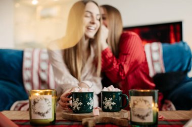 İki genç kız kardeş ellerinde yeşil bardakta marşmelov, neşeli, rahat bir atmosfer, Noel havası, kırmızı, yeşil renkler, masada mumlar.