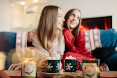 İki genç kız kardeş ellerinde yeşil bardakta marşmelov, neşeli, rahat bir atmosfer, Noel havası, kırmızı, yeşil renkler, masada mumlar.