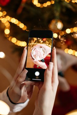 İki genç kız kardeş ellerinde marşmelovla yeşil fincan kakao tutuyorlar. Evlerinde neşeli, rahat bir atmosferdeler. Noel ağacının yanında birbirlerinin fotoğraflarını çekiyorlar.