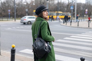 Yeşil elbiseli ve şapkalı bir kız otobüs durağında durmuş, sırtından otobüs manzarasını bekliyor.