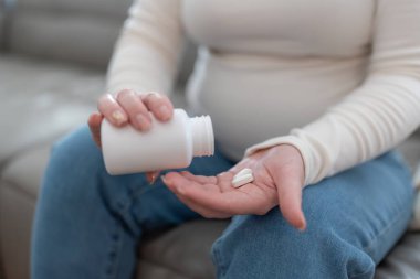 Tanımlanamayan hamile bir kadın elinde plastik kavanozla elinde haplar, vitamin ve mineraller tutarak evdeki koltukta oturup hamilelik boyunca hastalıkları tedavi ediyor. Yüksek kalite fotoğraf