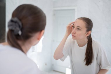 Mutsuz gen z kadın alnında bir sivilce buluyor, bir aynanın önünde duruyor. Sivilce tedavisi, cilt bakımı, yetişkin sivilcesi. Yüksek kalite fotoğraf